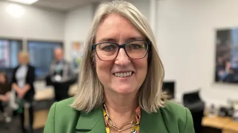 Labour skills minister Jacqui Smith stands in front of students at Dudley College - she has shoulder length blond hair, and is wearing glasses and a green jacket