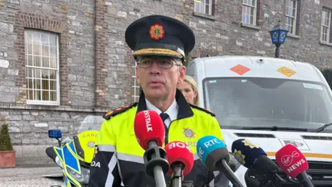 PA Media Garda Commissioner Drew Harris, wearing his Garda uniform, standing in front of a number of media microphones. There is a white van and a grey brick building behind him. 