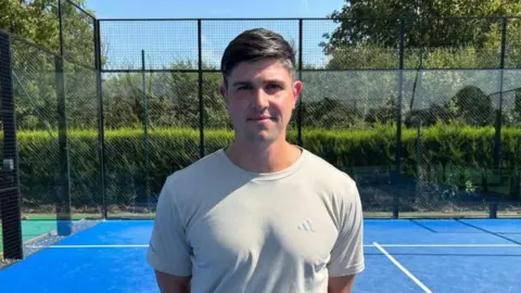 Charlie Stubbs / BBC There is a man in a white adidas t-shirt. He has black-brown hair which is brushed to one side. He is on a blue padel court. Behind him is the glass walls which make up the court.