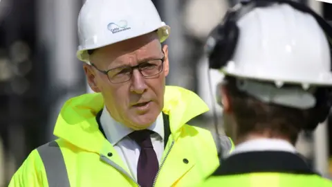 John Swinney, wearing glasses, a hard hat and high-visibility jacket, speaks to someone with dark hair in the foreground 