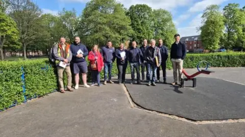Walsall Council Ten people are lined up against a hedge in Leamore Park, with a piece of playground equipment to the right of the group.