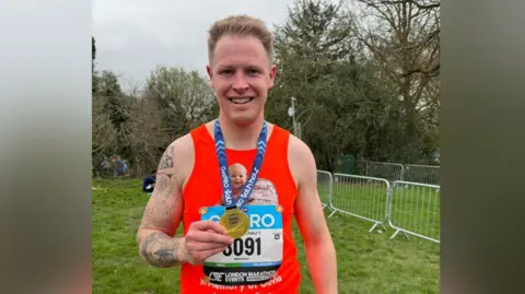 Harry Foster Harry Foster is wearing an orange running vest while holding a gold medal around his neck. He is posing with it while smiling.