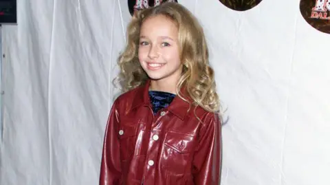 Getty Images Panettiere pictured smiling at 11-years-old at a film premiere. She has curly dark blonde hair and wears a vinyl dark red jacket with a crused velvet blue top.
