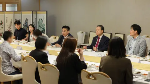 Men in suits and women in blazers seated at a long table, with meals laid out of each person. 
