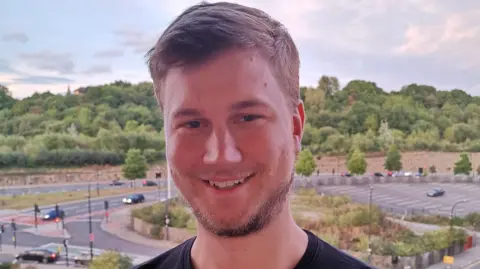 A man with short brown hair and stubble smiles at the camera. He is wearing a black T-shirt and standing with a road junction in the background