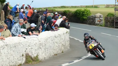 SOUTHERN 100 Alan Oversby racing at the event, he is on a black and orange machine with the number tow on the front on a yellow plate. He is rounding a bend while spectators watch on from behind a white-painted stone wall.