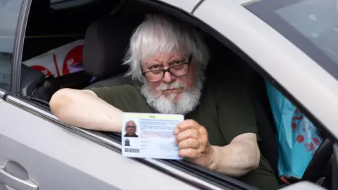 Pensioner Ralph James holding his Blue Badge while sitting in his car