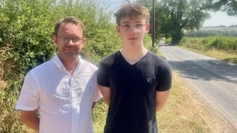 A man with a white shirt, stood next to his son, wearing a black t shirt. Both have short brown hair and glasses. They are stood on a grass verge next to a country lane.