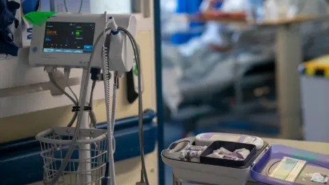 PA Media A monitor with wires hanging from it and a medical kit on a tray are in the foregoing of a photograph in a hospital. A patient in a bed is in the background.