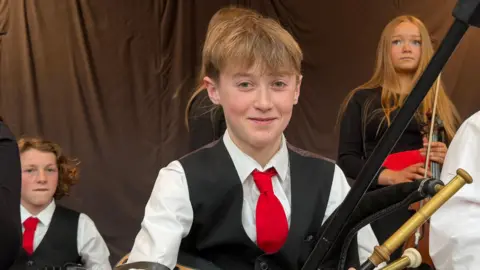 A boy with short blonde hair, wearing a white shirt, red tie and black waistcoat. There is a boy and a girl sat behind him. He is holding uilleann pipes.