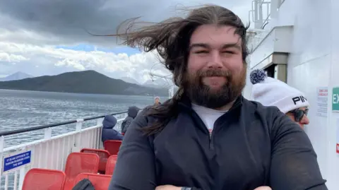 Martin is pictured outside on a ferry. His long dark hair is blowing in the wind, Martin, who has a full beard, is smiling and has his arms folded. There are other ferry passengers behind him, some of them sitting on red metal seats.