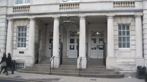 The exterior of a grey stone building. The image shows the steps and large pillar leading to three white doors.