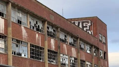The side of a derelict factory building, spanning multiple floors. There are numerous broken windows. There is graffiti on one of the upper sections of the building.