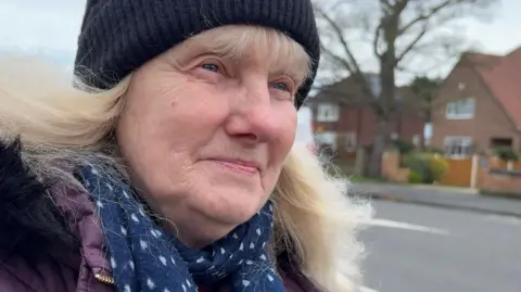 A woman with blond hair has a half-smile while looking into the middle-distance as she stands beside a road in a residential area. She is wearing winter clothes, including a black woolly hat, a purple coat and a blue scarf with white love heart design.