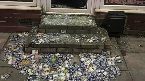 Celestial Collectibles Front step of a shop shows hundreds of Pokemon cards strewn on the ground mixed with smashed glass from the front door 