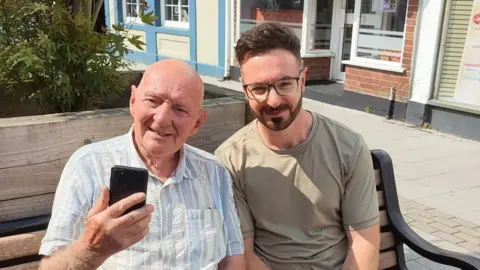 Comfort Companions CIC Grant Mackenzie, a man in his 30s with glasses and a beard, is sitting on a bench with his dad, who is wearing a stripy shirt and holding up a phone.