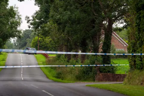 Pacemaker A blue and white police tape across a road with white marking in the middle. Grass verges and trees line the road. In the distance some cars a parked beside a red fence. 