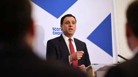 PA Media Anas Sarwar on stage at his media conference. He is standing in front of a large Saltire on the wall, faeturing the word Scotland's Choice in blue.