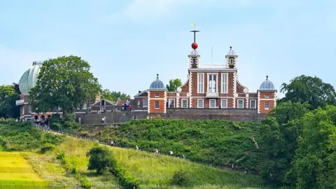 A 17th century building sits on top of a grassy hill surrounded by mature trees. There is a green circular building to the side that houses a telescope. 
