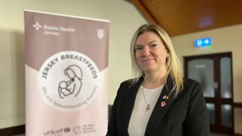 Raluca looks at the camera with a a sign behind her to her left which has the Breastfeeding Jersey Initiative logo on it. She is wearing a black jacket and has shoulder length blonde hair.