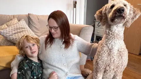 Picture shows a boy in army coloured t-shirt with blond hair cuddling his Mum who has long red hair and wears glasses - they're sat on the floor with a light coloured curly haired tall dog who is looking at the camera. 