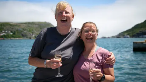 Cathie and Tricia pictured holding a glass of champagne each. They are both smiling. Tricia has her arm around Cathie.