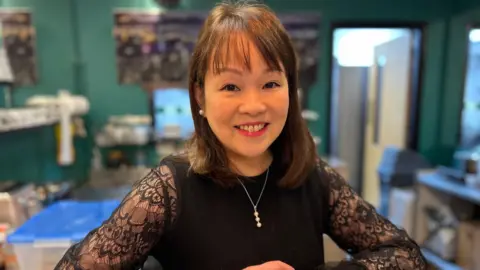 A woman in a black top with lacey sleeves smiling at a counter. She has black hair and is wearing a silver necklace and pearl earrings. Behind her a commercial kitchen area can be seen out of focus.