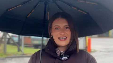 Woman with umbrella, mid speaking. She is wearing a brown jacket and is holding a black umbrella over her head.