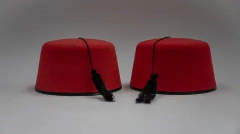 Nathan Fitzsimmons Two red fezzes, rimless hats with black tassels, photographed on a plain white background.