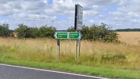Green road sign at the side of the A15 directing people either towards Sleaford or Lincoln. There is a section of road in view, along with a hedge and a field in the background.
