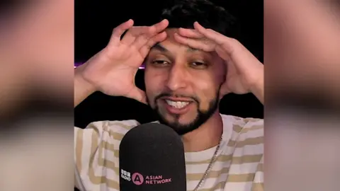 A man with short, dark hair and a short, dark beard holds his hands on his head as he speaks into a microphone.