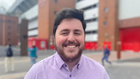 James Barber smiles while standing in front of Anfield Stadium. He is wearing a light purple shirt