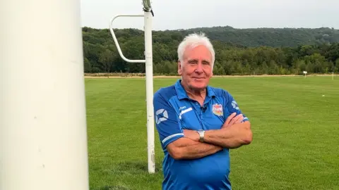 James Vincent / BBC Martin Windle d'Ecclesall Rangers regarde la caméra avec ses bras pliés. Il a les cheveux blancs et porte un maillot de football bleu. Il se trouve entre 2 éléments cibles - dont l'un est au premier plan de l'image.