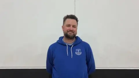 An image of Joe Hind, coach of the Guernsey Dynamics Football Club, in a blue Everton Football Club hoodie standing in front of a white wall smiling. 