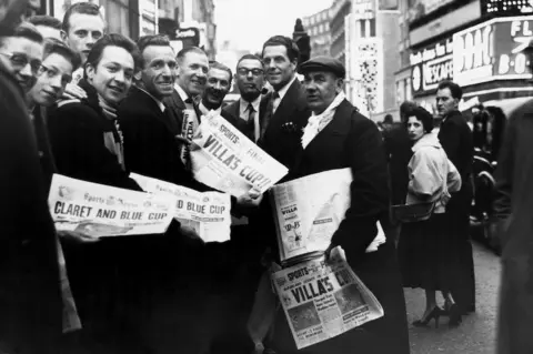 Getty Images A black and white image of football fans holding up newspapers, including the Sports Argus, with headlines about Aston Villa's win against Manchester United. 