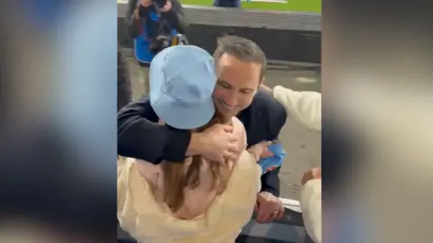 A young woman in a sky blue hat being hugged by a football manager in a black jacket