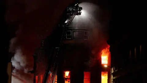 Getty Images A fire raging in a tall building, with a fireman on a extendable ladder spraying water over it