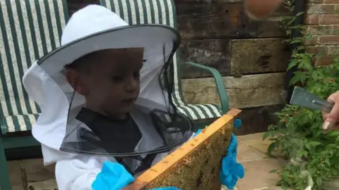 Supplied A child wearing a white beekeeping suit. He is stood in a garden holding a hive.