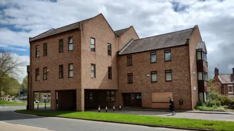 Large multi-storey brick building with several windows. A set of windows on the bottom floor are boarded up. There are two people walking past the building.