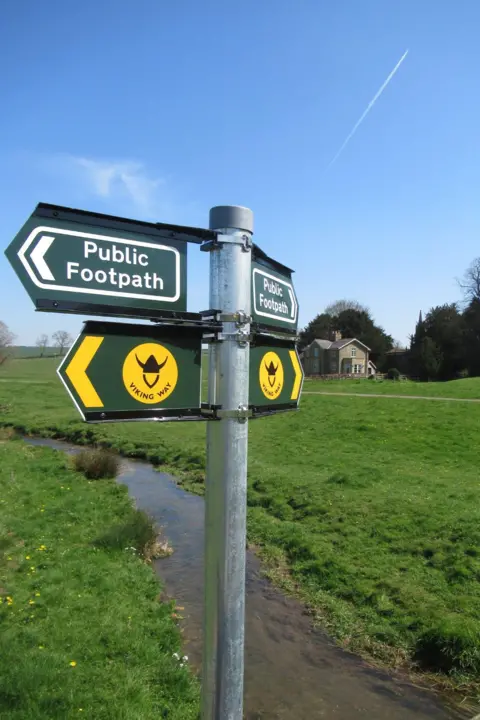 Lincolnshire County Council A pole with public footpath signs and black/yellow Viking helmet signs. There is a stream behind it surrounded by grass, and a house in the distance.