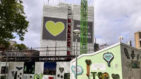 An image of the partly deconstructed tower surrounded by hoardings. There are tributes painted on to and attached to the hoardings.