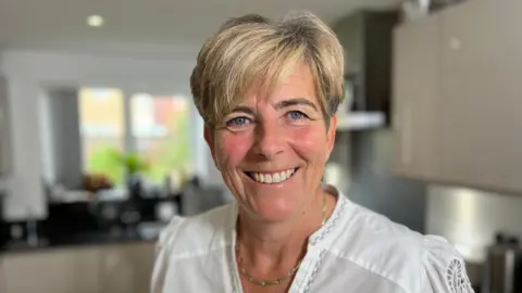 Cancer survivor Lisa Noble with short blonde highlighted hair and blue eyes. She is wearing a white top and gold necklace. She is smiling into the camera while standing in a kitchen, which is blurred in the background.