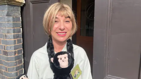 A woman with blonde hair wearing a white shirt smiles at the camera outside a building, with two toy monkeys draped around her neck and a small book badge pinned to her chest.