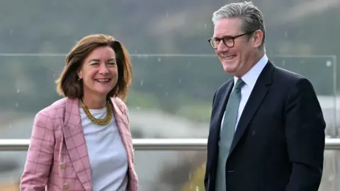 PA Media Eluned Morgan and Sir Keir Starmer stood next to each other with a steel bar behind them. Morgan is wearing a pink jacket and white top while Sir Keir is wearing a black suit, white shirt and green tie.