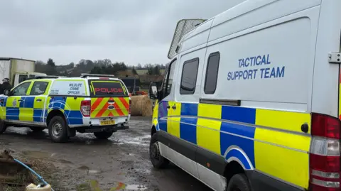 Cambridgeshire Police Two police tactical support vehicles are parked on what looks to be a farm. The ground is muddy with some puddles and there are fields and hay bales around them.