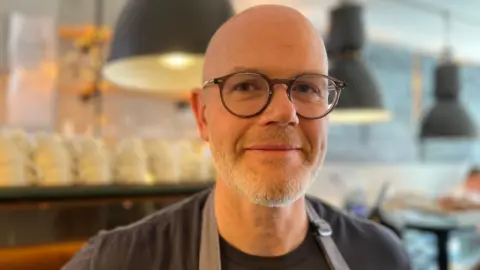 Man with bald head and glasses wearing apron stands in front of coffee machine