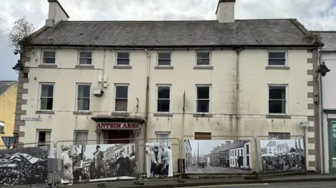 The former Antrim Arms hotel in Ballycastle town centre in September 2025. The large cream-coloured building has six front windows on each of its three floors and the sign above the front door says "Antrim Arms" in red lettering. There are two chimneys and vegetation is growing out of the guttering at both sides of the roof. Two of the front windows are boarded up. Steel safety barriers, decorated with old photos of the town, stand in front of the building. 