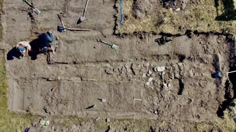Supplied Overhead aerial view of an archaeological excavation site showing Meg Russell and Dr Derek Pitman working within a rectangular trench. Exposed stone features, soil layers, and several excavation tools are visible across the dig area, which is bordered by grass and marked excavation edges.