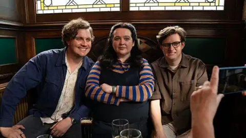 Two Brothers Productions Ashley Storrie as Nina in a scene from Dinosaur. She is wearing a striped blue and orange top and is sitting in-between Clay - wearing a white patterned shirt and denim jacket - and Lee, who is smiling awkwardly while wearing a brown shirt and white jeans. He has glasses on. The trio are sitting in a pub, and someone is taking a photo of them on a phone.