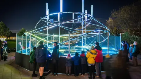 Barnsley council Around 20 people are standing around the edge of a large art installation made up of brightly lit tubes twisting around each other in a loose, haphazard spiral shape.